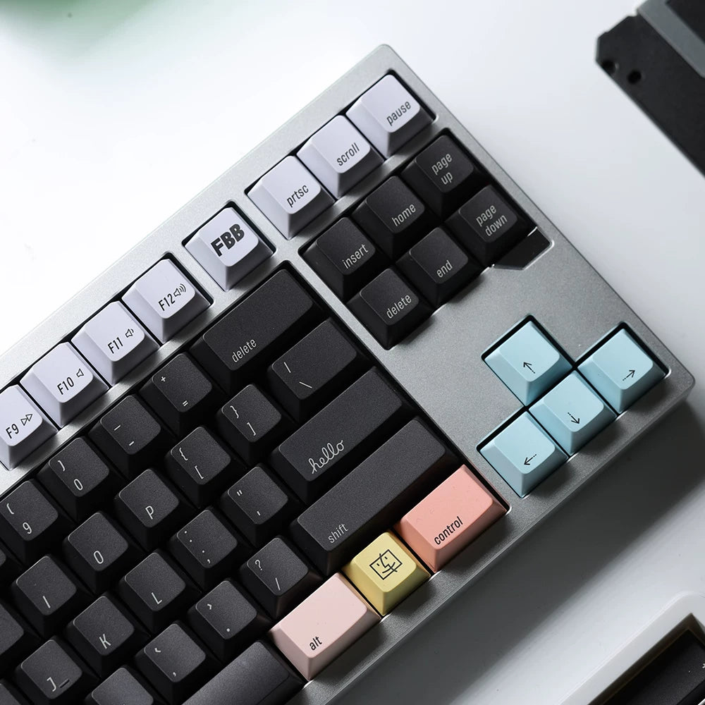 A close-up of a mechanical keyboard with a silver-gray chassis, black keycaps, and pastel-colored accent keys including light blue, peach, pink, and gold. The keyboard is photographed at an angle on a clean white surface.