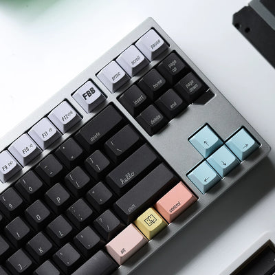 A close-up of a mechanical keyboard with a silver-gray chassis, black keycaps, and pastel-colored accent keys including light blue, peach, pink, and gold. The keyboard is photographed at an angle on a clean white surface.