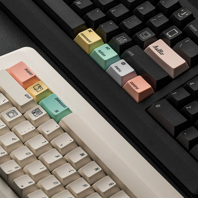 Close-up of a mechanical keyboard with custom pastel keycaps. The keyboard features a cream-colored main body with peach, yellow, and mint green accent keycaps, alongside a black keyboard in the background with peach, yellow, mint green, gray, and beige accent keycaps.